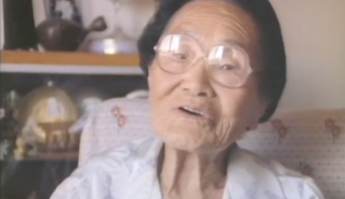Haruo Kasahara sings ho-le-ho-le bushi for the camera sitting on an upholstered chair with a bookshelf in the background